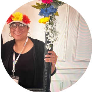 Elsa Johnson headshot. She has medium light skin and is wearing all black and a hat with flowers on top. She's posing behind a flowery picture frame.